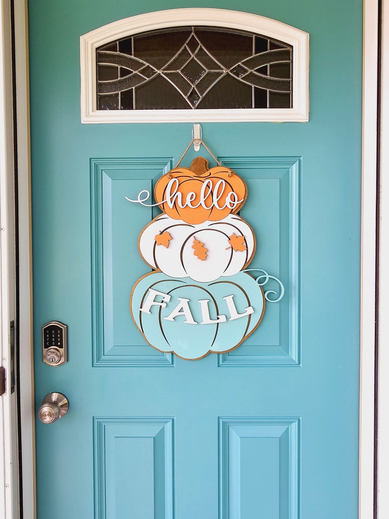 Op de afbeelding: Een turquoise voordeur met een houten pompoenkrans waarop "hello fall" staat in witte en oranje letters.