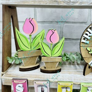 May include: Two wooden tulips painted in shades of pink and green, with brown pots, sitting on a shelf with a wooden sign that says "Spring" in colorful letters.