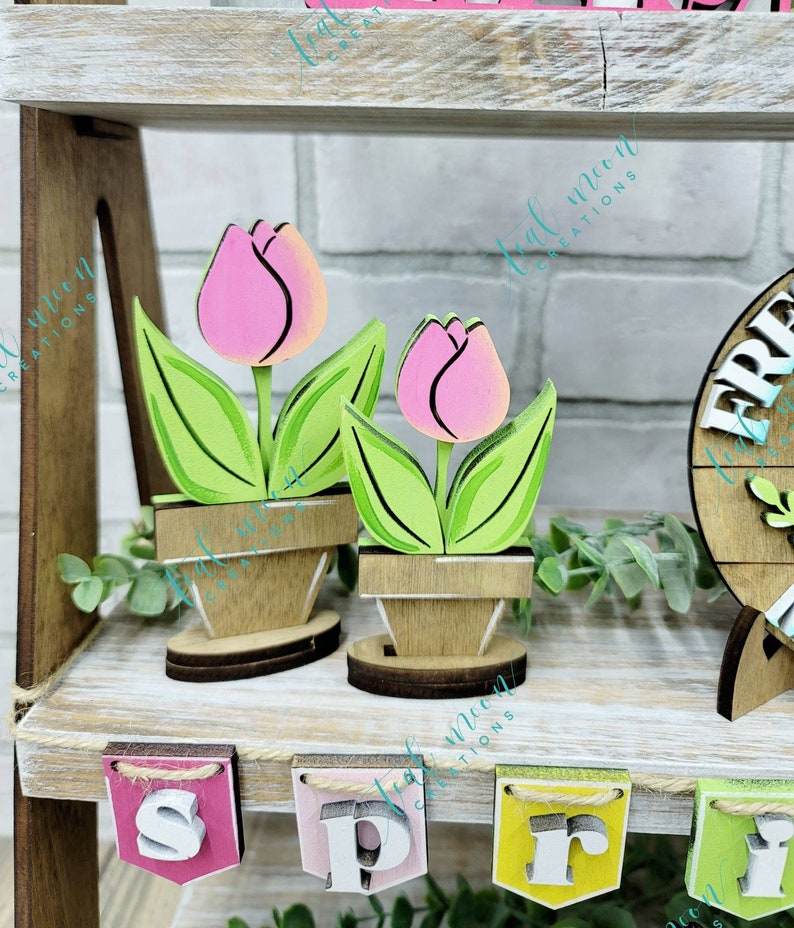 May include: Two wooden tulips painted in shades of pink and green, with brown pots, sitting on a shelf with a wooden sign that says "Spring" in colorful letters.
