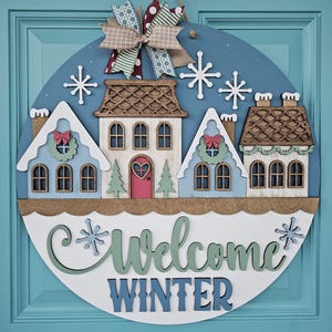 May include: A round wooden winter-themed door sign with a blue background and a decorative bow. The sign features three houses, snowflakes, and the words "Welcome Winter" in a script font. The houses are painted in white, blue, and brown.
