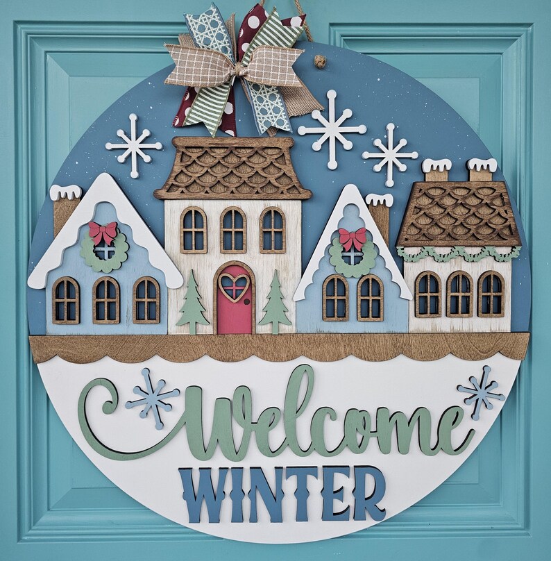 May include: A round wooden winter-themed door sign with a blue background and a decorative bow. The sign features three houses, snowflakes, and the words "Welcome Winter" in a script font. The houses are painted in white, blue, and brown.