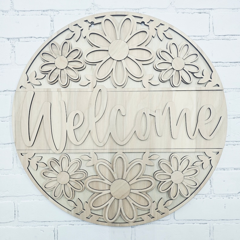 K&ouml;nnte beinhalten: Ein rundes Holzschild mit einer Willkommens-Botschaft in kursiver Schrift. Das Schild ist mit einem floralen Design verziert, das mehrere g&auml;nsebl&uuml;mchenartige Blumen zeigt.