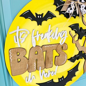 Op de afbeelding: Een ronde gele deurhanger met de tekst "It's Freaking Bats in Here!" in witte en bruine letters. De deurhanger is versierd met zwarte vleermuizen.