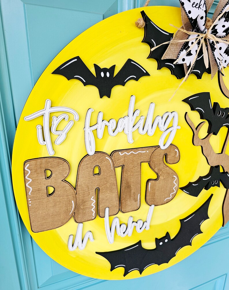 Op de afbeelding: Een ronde gele deurhanger met de tekst "It's Freaking Bats in Here!" in witte en bruine letters. De deurhanger is versierd met zwarte vleermuizen.