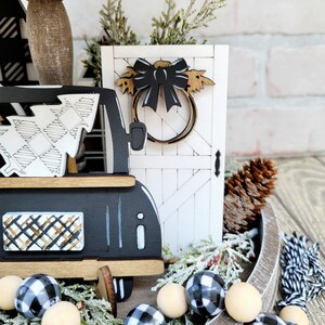 May include: A tiered tray decorated with a black and white plaid truck, a white wooden door with a black bow and wreath, a pine cone, and a black and white plaid garland with wooden beads.