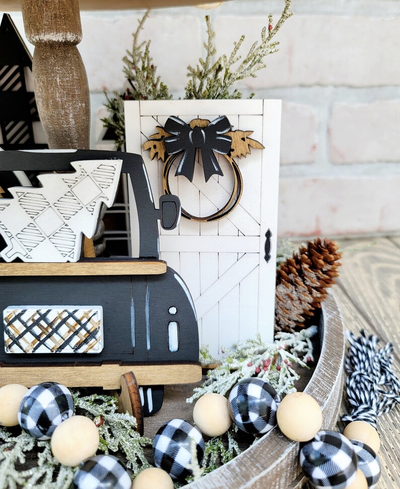 May include: A tiered tray decorated with a black and white plaid truck, a white wooden door with a black bow and wreath, a pine cone, and a black and white plaid garland with wooden beads.
