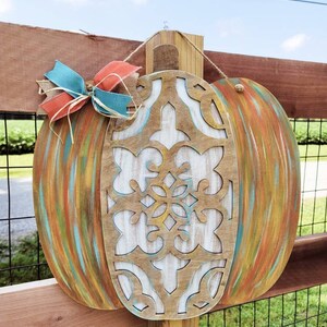 May include: A wooden pumpkin door hanger with a carved design and painted in shades of blue, green, orange, and brown. The pumpkin is decorated with a burlap bow and a red and blue ribbon.