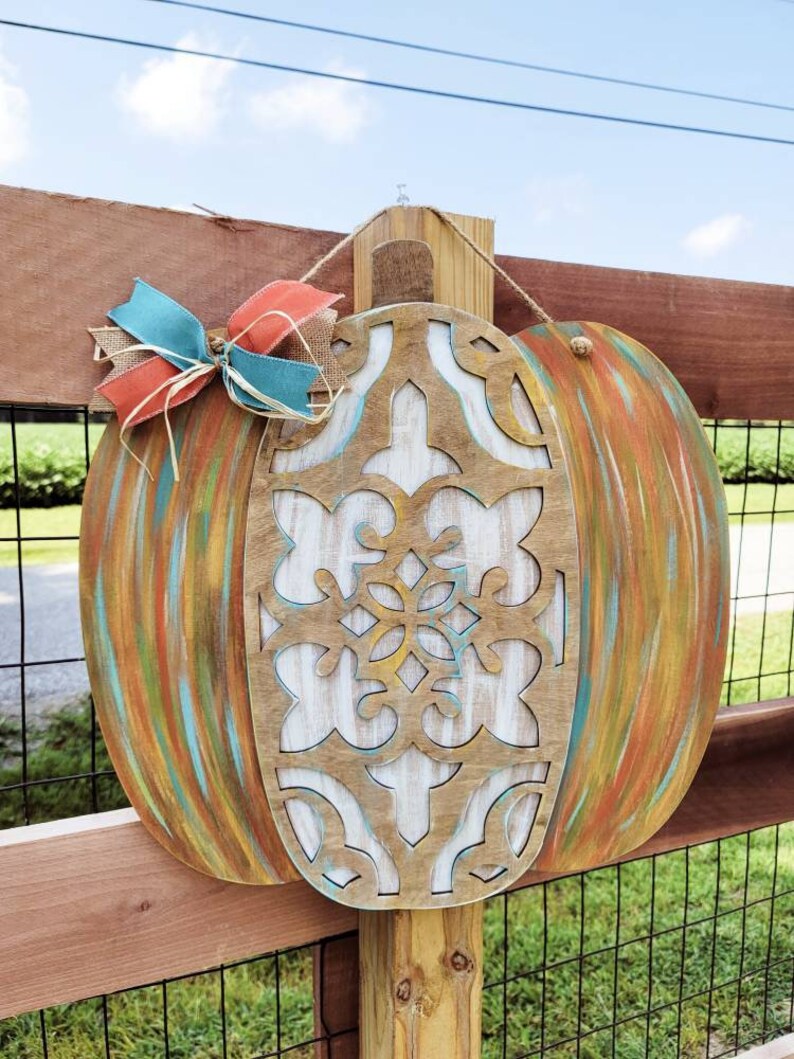 May include: A wooden pumpkin door hanger with a carved design and painted in shades of blue, green, orange, and brown. The pumpkin is decorated with a burlap bow and a red and blue ribbon.