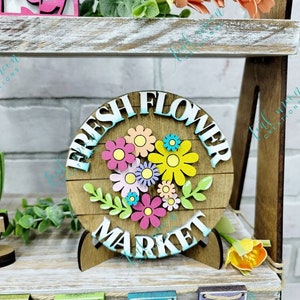 May include: A wooden sign with the words "Fresh Flower Market" in white letters. The sign is decorated with colorful flowers and green leaves. The sign is on a wooden stand.
