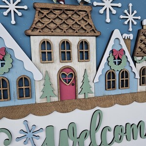 May include: A wooden winter-themed welcome sign featuring a village scene with houses, snowflakes, and the words "Welcome Winter." The houses are painted in shades of blue, white, and brown, with a pink door and green trees. The sign is decorated with a bow.