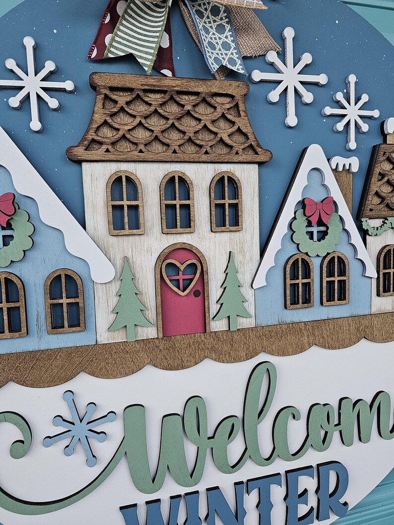 May include: A wooden winter-themed welcome sign featuring a village scene with houses, snowflakes, and the words "Welcome Winter." The houses are painted in shades of blue, white, and brown, with a pink door and green trees. The sign is decorated with a bow.
