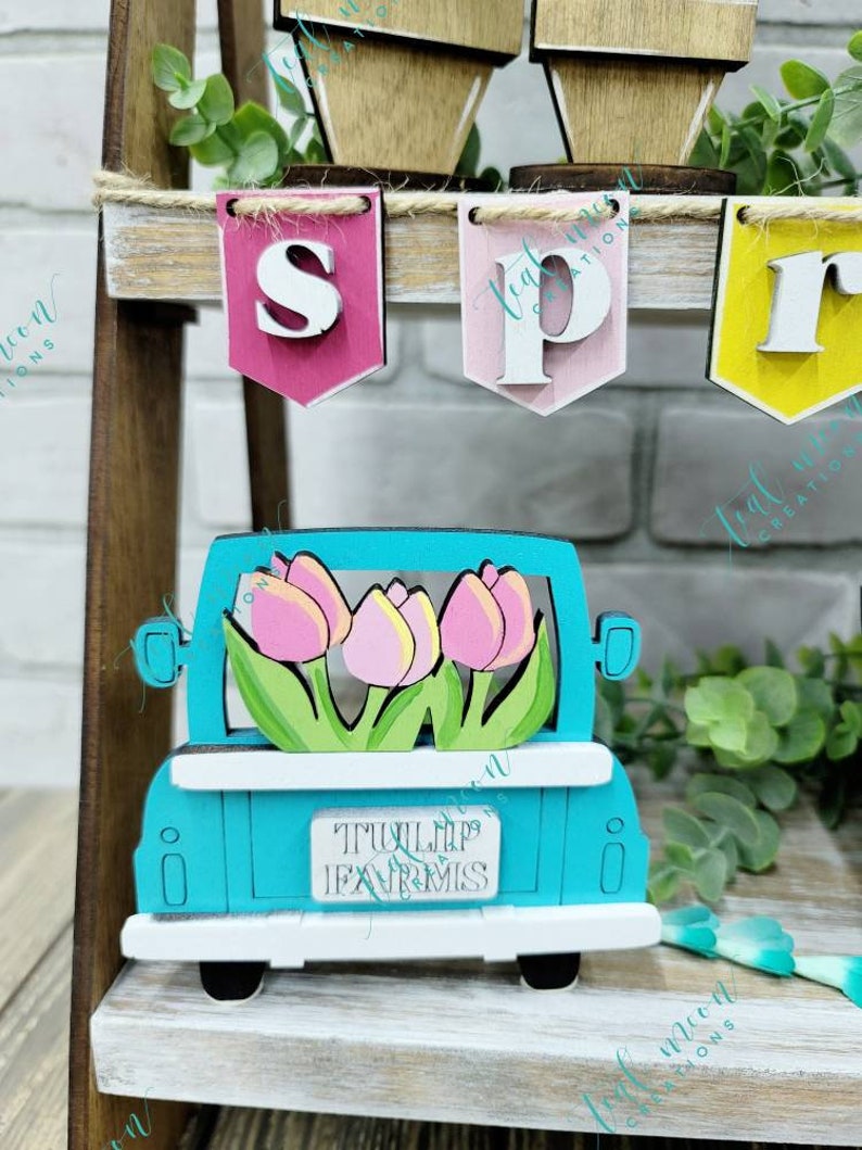 May include: A teal blue wooden truck with a white bed and white wheels. The truck is carrying a bouquet of pink and yellow tulips. The truck has a sign that reads "TULIP FARMS".