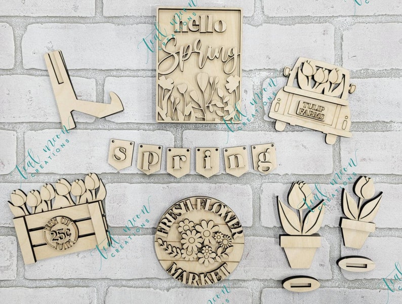May include: A set of wooden spring-themed craft supplies, including a sign that says "hello spring", a truck with tulips, a flower market sign, a flower pot, and a banner that says "spring".