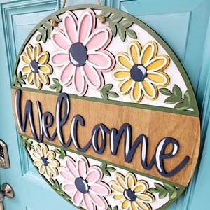K&ouml;nnte beinhalten: Ein rundes, h&ouml;lzernes Willkommensschild mit wei&szlig;em Hintergrund und gr&uuml;nem Rand. Das Schild zeigt ein Blumenmuster mit rosa und gelben Blumen und gr&uuml;nen Bl&auml;ttern. Das Wort "Willkommen" ist in dunkelblauer, kursiv geschriebener Schrift geschrieben.