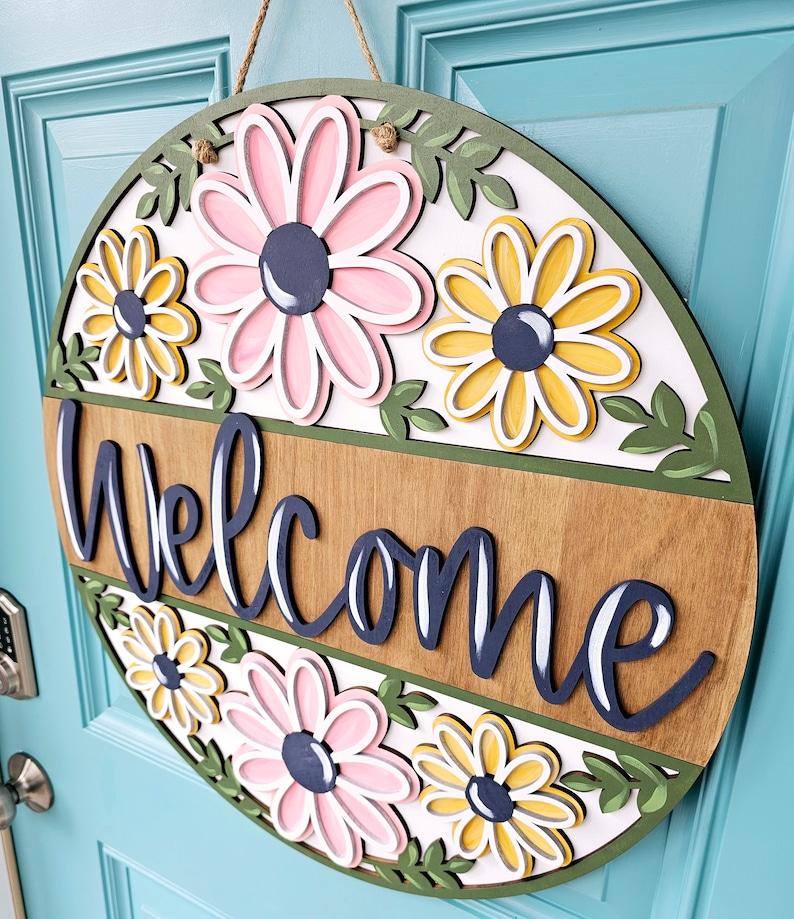 K&ouml;nnte beinhalten: Ein rundes, h&ouml;lzernes Willkommensschild mit wei&szlig;em Hintergrund und gr&uuml;nem Rand. Das Schild zeigt ein Blumenmuster mit rosa und gelben Blumen und gr&uuml;nen Bl&auml;ttern. Das Wort "Willkommen" ist in dunkelblauer, kursiv geschriebener Schrift geschrieben.