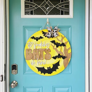 Op de afbeelding: Een gele en bruine Halloween-deurkrans met de tekst "It's Freaking Bats in Here!" en zwarte vleermuizen. De krans hangt aan een teal blauwe deur.