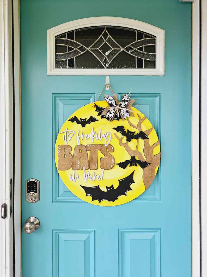 Op de afbeelding: Een gele en bruine Halloween-deurkrans met de tekst "It's Freaking Bats in Here!" en zwarte vleermuizen. De krans hangt aan een teal blauwe deur.