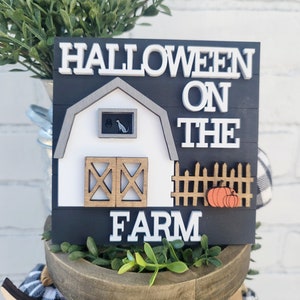 May include: A black and white wooden sign with the text "Halloween on the Farm". The sign features a white barn with a black cat in the window, a brown fence with two orange pumpkins, and a black background.