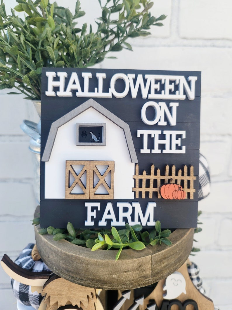 May include: A black and white wooden sign with the text "Halloween on the Farm". The sign features a white barn with a black cat in the window, a brown fence with two orange pumpkins, and a black background.