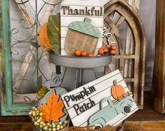 Könnte beinhalten: Ein zweistöckiger Metallständer mit grauer Oberfläche. Die obere Ebene zeigt ein Holzschild mit dem Wort "Thankful" und eine Holzeichel. Die untere Ebene zeigt ein Holzschild mit den Worten "Pumpkin Patch" und einen blauen LKW mit einem Kürbis in der Ladefläche. Der Ständer ist mit herbstlichen Dekorationen geschmückt, darunter ein Herbstblatt, ein kleiner Kürbis und Beeren.