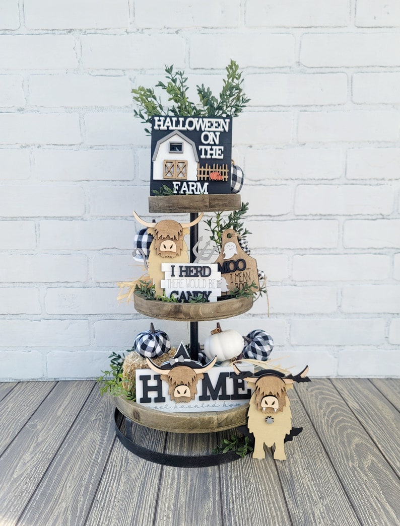 May include: A three-tiered wooden tray stand with black metal accents. The top tier features a black and white sign that says "Halloween on the Farm" with a small white farmhouse cutout. The middle tier has a black and white checkered pumpkin, a white pumpkin, a brown and white Highland cow cutout, and a sign that says "I herd there would be candy" and another sign that says "Moo I mean M". The bottom tier has a sign that says "Home" with a brown and white Highland cow cutout, a black and white checkered pumpkin, and a white pumpkin.