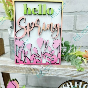 May include: A wooden sign with the words "hello spring" in a pink and green ombre gradient. The sign is decorated with a floral design and has a white background.