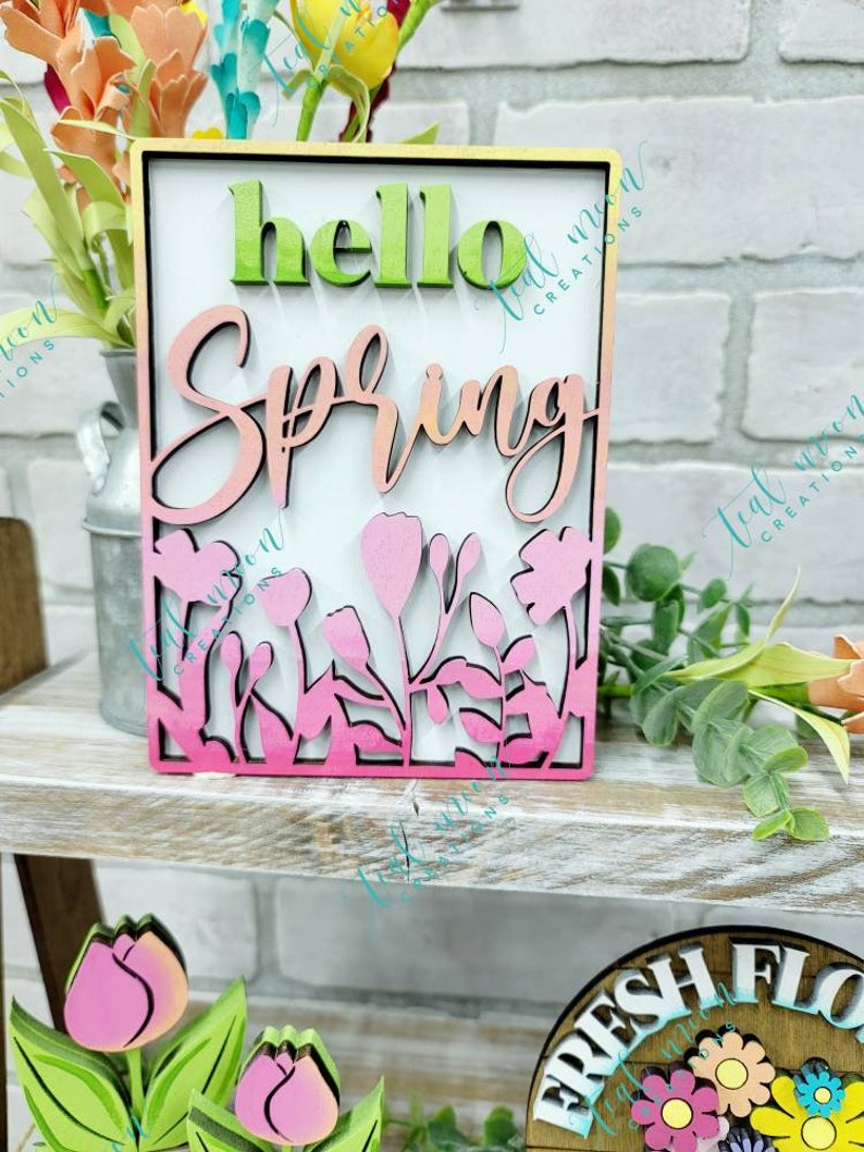 May include: A wooden sign with the words "hello spring" in a pink and green ombre gradient. The sign is decorated with a floral design and has a white background.