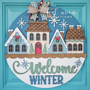 May include: A round wooden winter-themed door sign with a blue background, featuring three houses, snowflakes, and the words "Welcome Winter." The sign is adorned with a decorative bow and is designed for seasonal home decor.