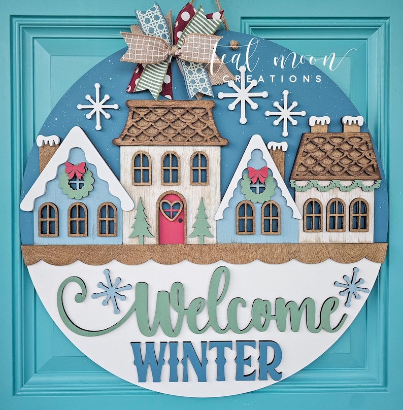 May include: A round wooden winter-themed door sign with a blue background, featuring three houses, snowflakes, and the words "Welcome Winter." The sign is adorned with a decorative bow and is designed for seasonal home decor.