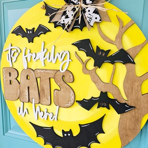 Op de afbeelding: Een ronde, gele, houten deurhanger met de woorden "It's Freaking Bats in Here!" in witte en bruine letters. De deurhanger is versierd met zwarte vleermuizen en een bruine tak.