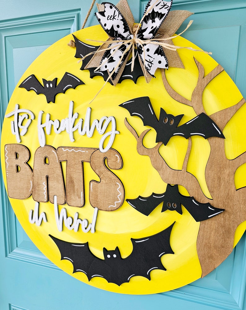 Op de afbeelding: Een ronde, gele, houten deurhanger met de woorden "It's Freaking Bats in Here!" in witte en bruine letters. De deurhanger is versierd met zwarte vleermuizen en een bruine tak.