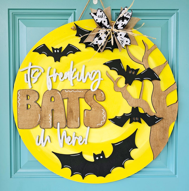 Op de afbeelding: Een ronde, gele, houten deurhanger met de tekst "It's Freaking Bats in Here!" in witte letters. De deurhanger is versierd met zwarte vleermuizen en een bruine tak.