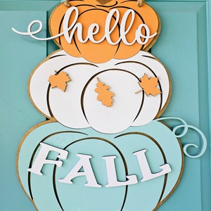Op de afbeelding: Een gelaagde pompoen deurhanger met de woorden "hello fall" in witte letters. De bovenste pompoen is oranje, de middelste pompoen is wit met oranje bladeren en de onderste pompoen is lichtblauw.