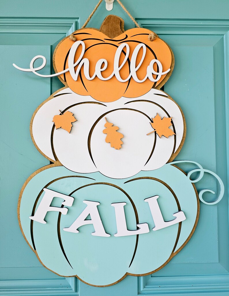 Op de afbeelding: Een gelaagde pompoen deurhanger met de woorden "hello fall" in witte letters. De bovenste pompoen is oranje, de middelste pompoen is wit met oranje bladeren en de onderste pompoen is lichtblauw.