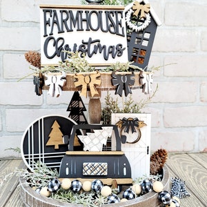May include: A tiered tray decorated for Christmas with a farmhouse theme. The tray features a black and white sign that says "Farmhouse Christmas", a black and white truck carrying a Christmas tree, a black and white door with a bow, and a black and white house with a wreath. The tray is decorated with black and white buffalo check ribbon, black and white buffalo check beads, and pine sprigs.