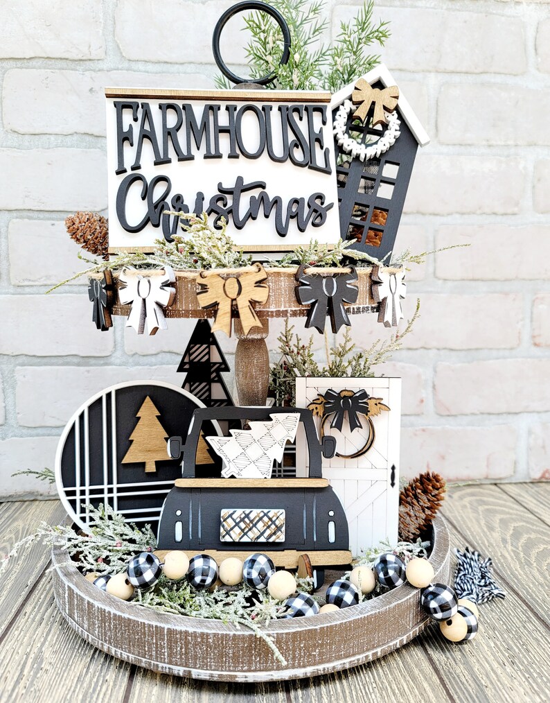May include: A tiered tray decorated for Christmas with a farmhouse theme. The tray features a black and white sign that says "Farmhouse Christmas", a black and white truck carrying a Christmas tree, a black and white door with a bow, and a black and white house with a wreath. The tray is decorated with black and white buffalo check ribbon, black and white buffalo check beads, and pine sprigs.