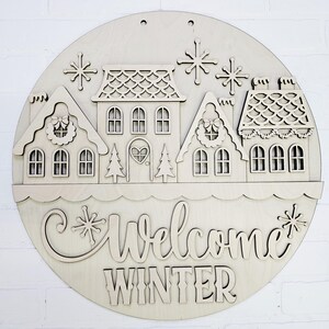 May include: A round wooden sign with a winter village scene. The sign features cut-out houses, snowflakes, and the words "Welcome Winter". The houses have intricate details, including wreaths and heart-shaped door. The sign is made of light-colored wood.