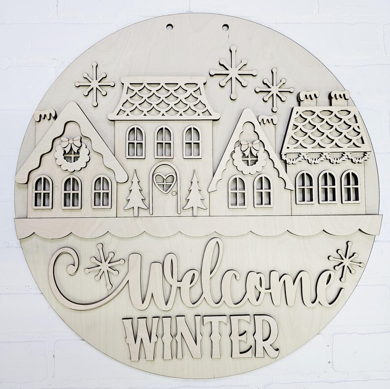 May include: A round wooden sign with a winter village scene. The sign features cut-out houses, snowflakes, and the words "Welcome Winter". The houses have intricate details, including wreaths and heart-shaped door. The sign is made of light-colored wood.