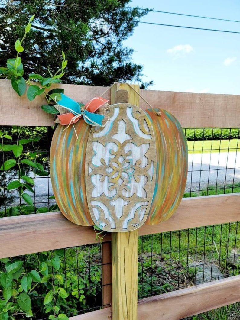 May include: A wooden pumpkin with a carved design painted in white, teal, and orange. The pumpkin is hanging on a wooden fence with a green vine growing on it.