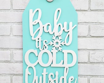 Può includere: Un cartello in legno blu turchese con un nastro e un fiocco bianchi appesi in alto. Il cartello ha la forma di un'etichetta e presenta il testo "Baby it's cold outside" scritto in lettere bianche in corsivo con un fiocco di neve tra le parole "it's" e "cold".