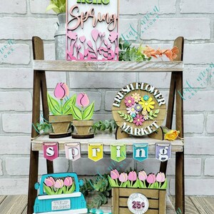 May include: A tiered wooden shelf display decorated with spring-themed wooden crafts. The top shelf features a sign that says "hello spring" with pink and green flowers. The middle shelf has a sign that says "fresh flower market" with a floral wreath and two potted tulips. The bottom shelf has a banner that says "spring", a blue truck with tulips in the back, and a wooden crate with tulips and a sign that says "25c".