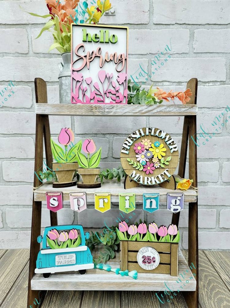 May include: A tiered wooden shelf display decorated with spring-themed wooden crafts. The top shelf features a sign that says "hello spring" with pink and green flowers. The middle shelf has a sign that says "fresh flower market" with a floral wreath and two potted tulips. The bottom shelf has a banner that says "spring", a blue truck with tulips in the back, and a wooden crate with tulips and a sign that says "25c".