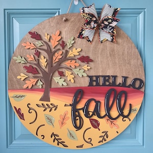 May include: A round wooden fall-themed door sign with a tree, leaves, and the words "HELLO FALL." The sign features a colorful design with orange, yellow, and red hues, and is adorned with a decorative bow.