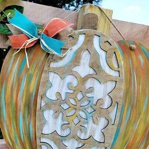 May include: A wooden pumpkin door hanger with a carved design and painted in shades of blue, green, yellow, and orange. The pumpkin is decorated with a turquoise and orange bow.
