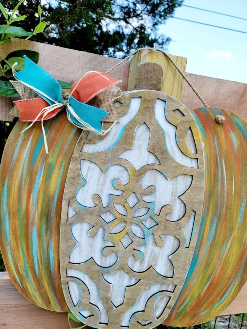 May include: A wooden pumpkin door hanger with a carved design and painted in shades of blue, green, yellow, and orange. The pumpkin is decorated with a turquoise and orange bow.