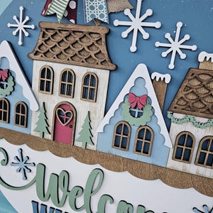 May include: A round wooden winter-themed sign with a blue background, featuring a village scene with houses, snowflakes, and the words "Welcome WINTER." The sign is adorned with a decorative bow at the top.