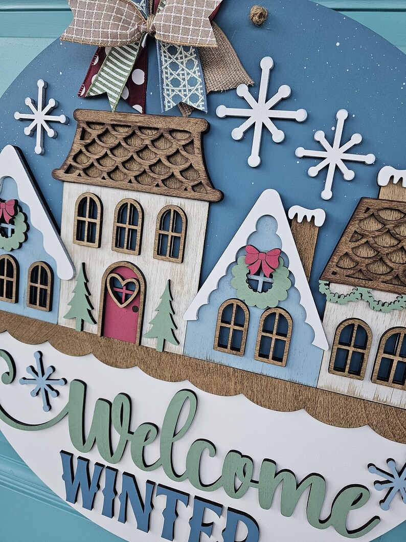 May include: A round wooden winter-themed sign with a blue background, featuring a village scene with houses, snowflakes, and the words "Welcome WINTER." The sign is adorned with a decorative bow at the top.