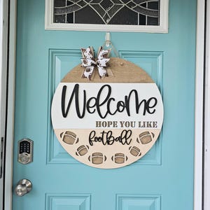 以下が含まれることがあります： 「Welcome Hope You Like Football」と書かれた丸い木製ドアハンガーと、茶色と白のリボン。背景は薄い青色のドアです。