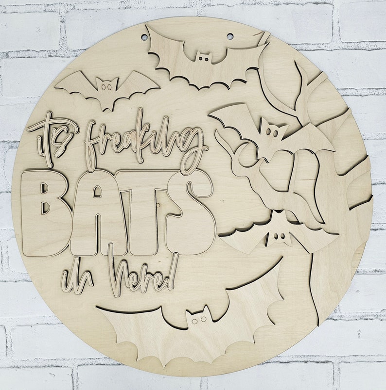 Op de afbeelding: Een rond houten bord met de tekst "It's Freaking Bats in Here!" en meerdere vleermuis uitsnijdingen. Het bord is geschilderd in een natuurlijke houtkleur.