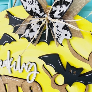 Op de afbeelding: Een gele houten deurhanger met de woorden "wreaking havoc" in wit schrift. De deurhanger is versierd met een zwart-witte vleermuis-thema strik, zwarte vleermuizen en bruine takken.